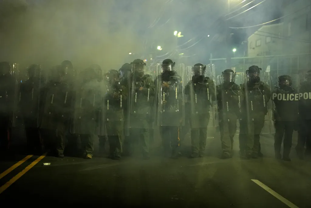 Law enforcement officers in riot gear stand in smoke during clashes with anti-ICE protesters.