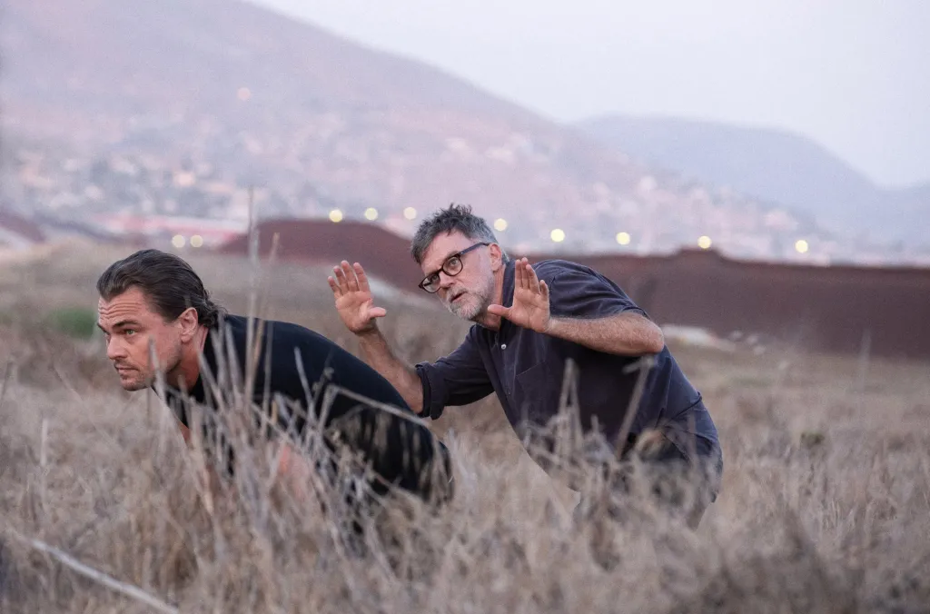 On set, Leonardo DiCaprio and director Paul Thomas Anderson crouch in dry grass, with Anderson holding his hands up.