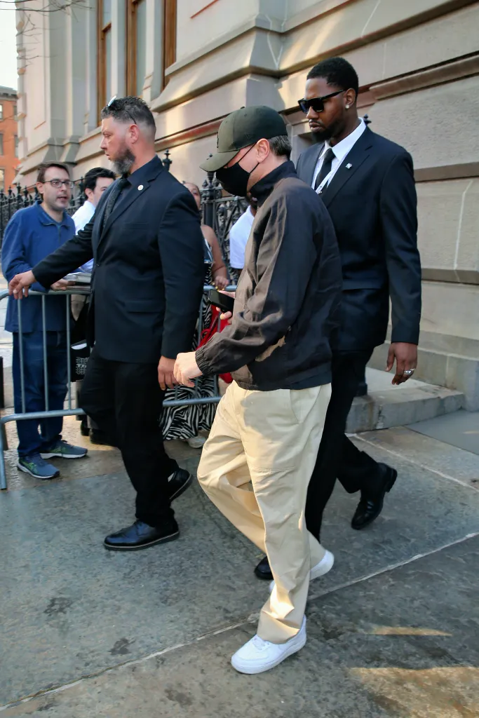 Leonardo DiCaprio walking, wearing a black cap and face mask, khaki pants, and white sneakers, accompanied by two bodyguards in suits.