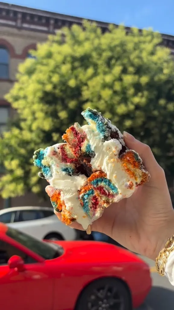 Hand holding a colorful donut with white frosting in front of a red car and trees.