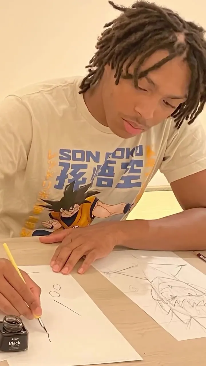 A man with dreadlocks wearing a Son Goku t-shirt is drawing with a pen and ink.