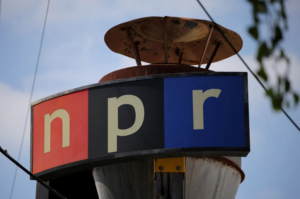 An outdoor sign with the NPR logo against a blue sky, with a rusted metal disk above it.