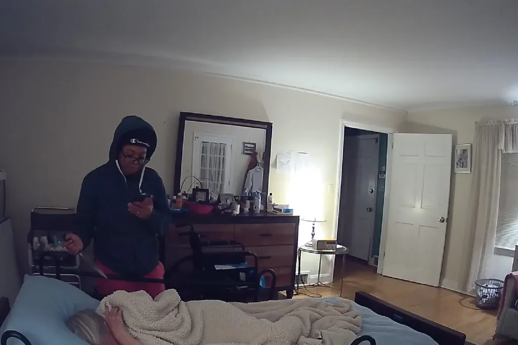 A person in a hoodie and glasses standing over an elderly person sleeping in a bed, holding a knife.