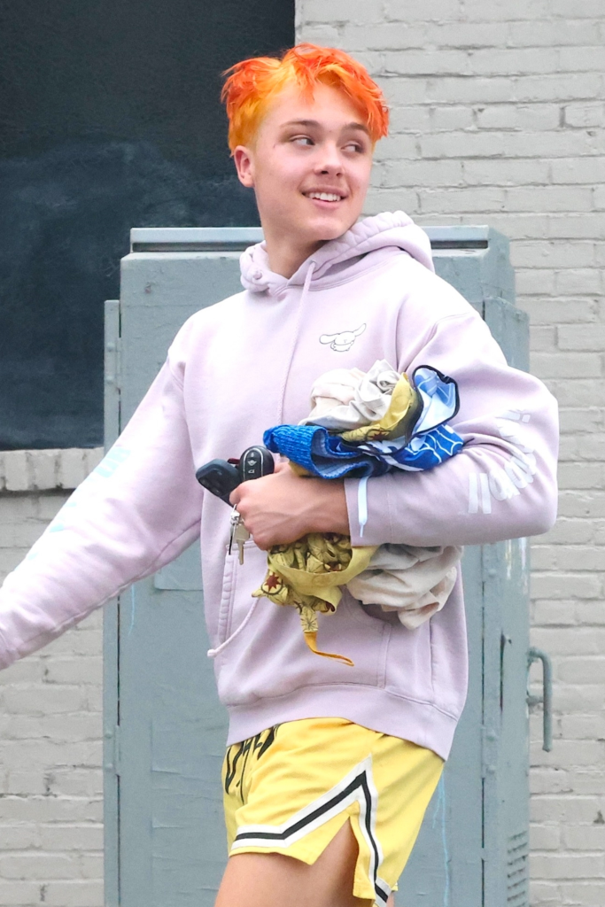 Knox Jolie-Pitt in a light purple hoodie and yellow shorts with bright orange hair, holding car keys and clothes.