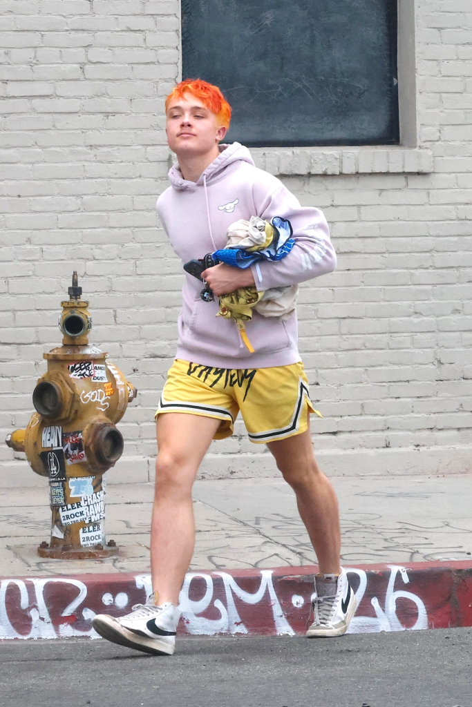 Knox Jolie-Pitt in yellow shorts and a purple hoodie, with bright orange hair, carrying clothes and keys, walking past a yellow fire hydrant and a white brick wall in Los Angeles.