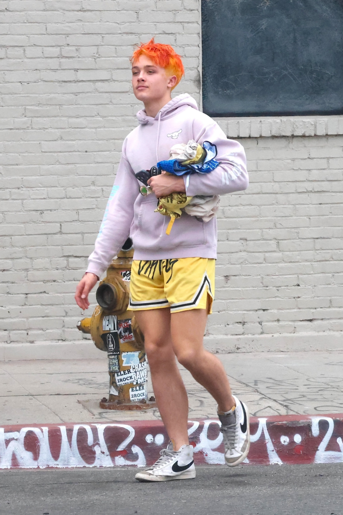 Knox Jolie-Pitt in a lavender hoodie, yellow shorts, and white Nike high-tops, walking with bright orange hair and holding items, possibly after a Muay Thai training session.
