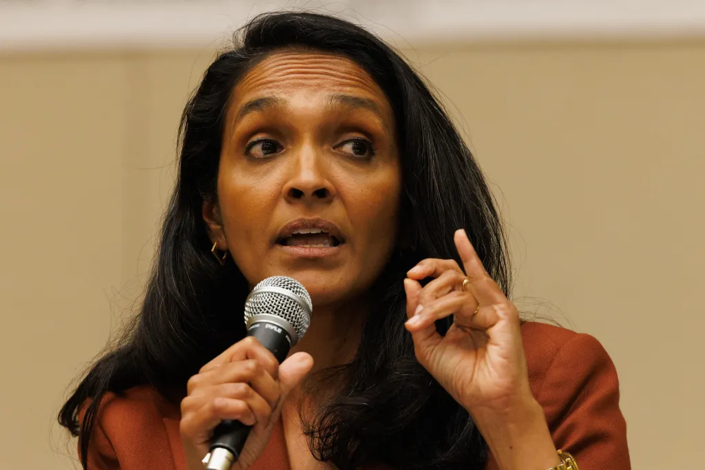 Nithya Raman speaking at a debate, holding a microphone in her left hand and gesturing with her right hand.