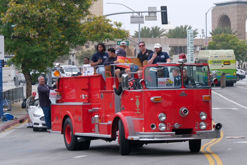 Firefighters on a vintage fire truck hold signs reading 