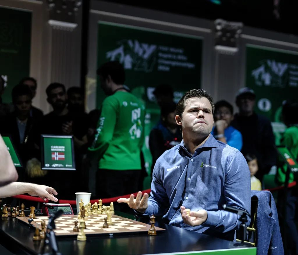 Magnus Carlsen playing chess, with his hands up in a gesture, possibly after a move or during a thought process, with the opponent's hand visible on the left and a chess clock showing 0.07.