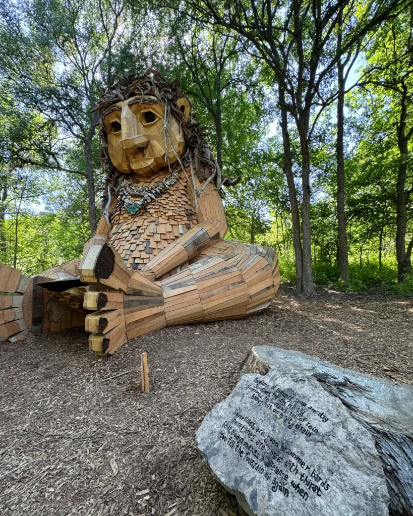 Malin's Fountain, a large wooden troll sculpture, sits in a forest next to a rock engraved with a poem.