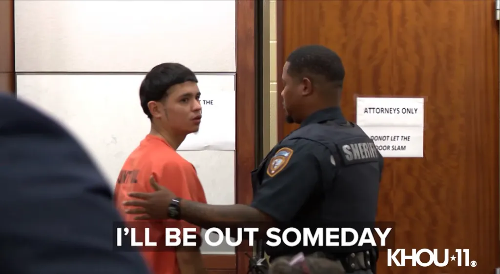 A man in an orange shirt being escorted by a sheriff in a courtroom.