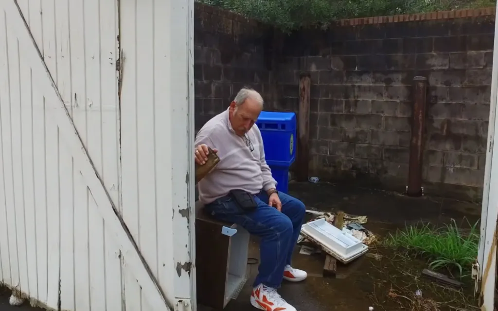 A man sits outside, looking down, next to a blue recycling bin and a brick wall.
