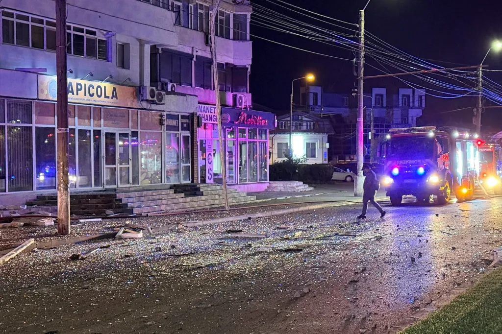 A man walks past scattered debris and glass on a street next to a building and fire trucks at night.