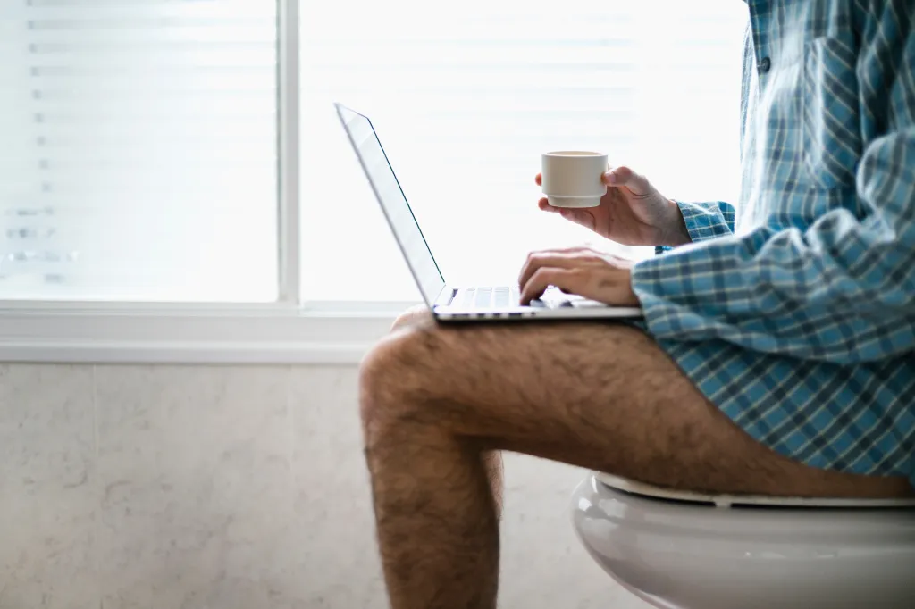 Man working on his laptop and holding a mug of coffee while sitting on the toilet at home, illustrating remote work.
