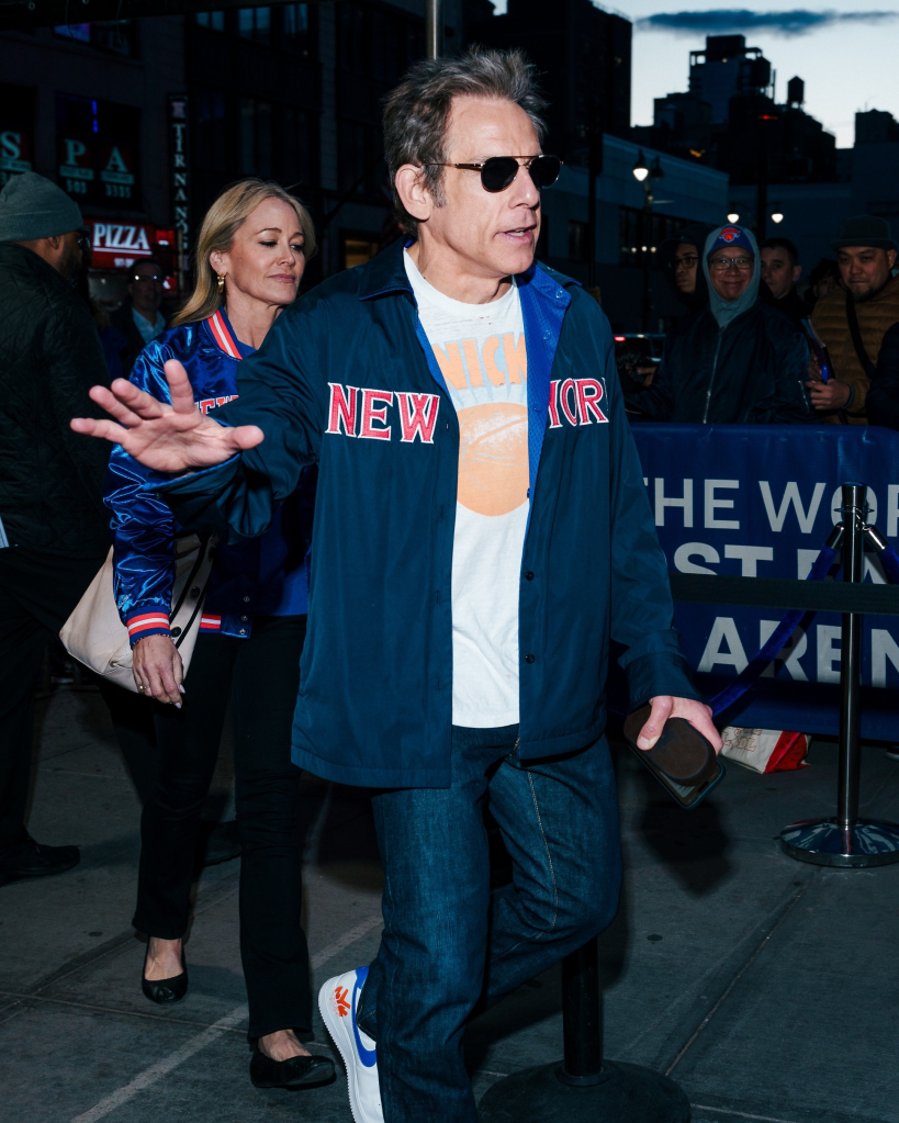 Ben Stiller wearing a blue New York Knicks jacket and sunglasses, with a woman beside him, walking past barriers at Game 2 of the Knicks playoff series.