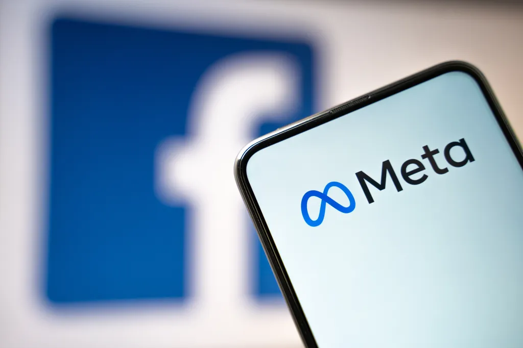 A smartphone displays the Meta logo, with a blurred Facebook logo in the background.