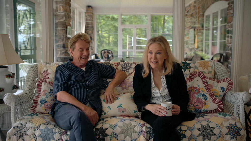 Martin Short and Catherine O'Hara sitting on a quilt-covered couch, both looking towards the right and smiling.