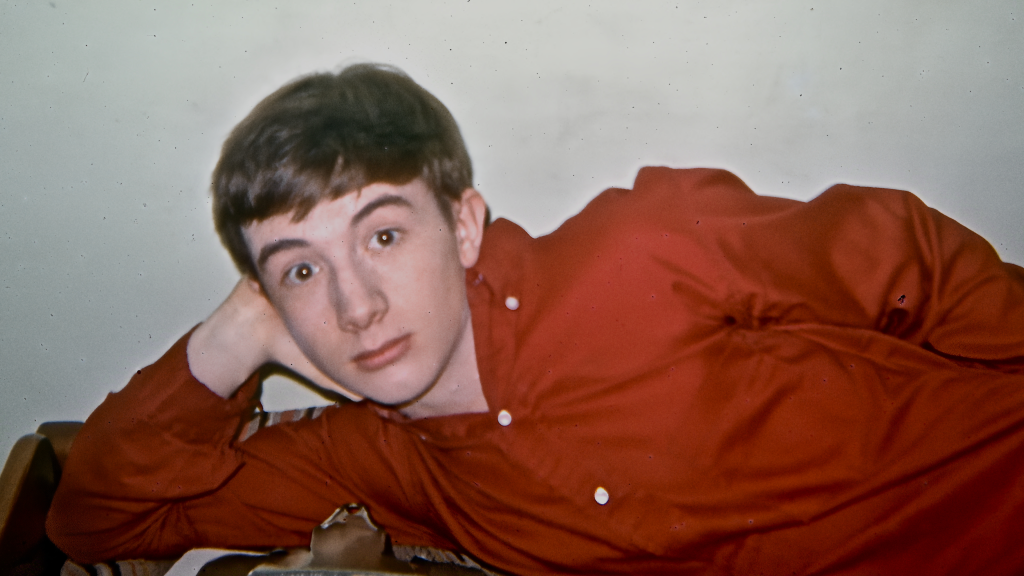 Martin Short as a young man with short brown hair, wearing a red shirt, lying down and resting his head on his hand.