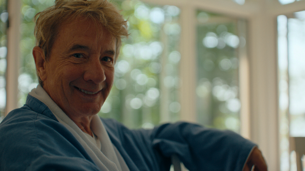 Martin Short in a blue robe, smiling at the camera.