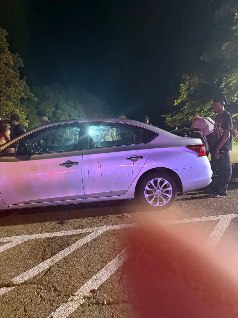 White car with bloodstains on its side in a parking lot at night, with people and trees in the background.