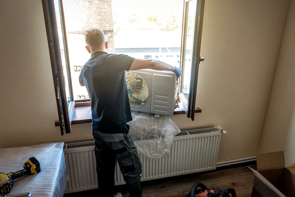 A technician installing the outer block of an air conditioner through a window.