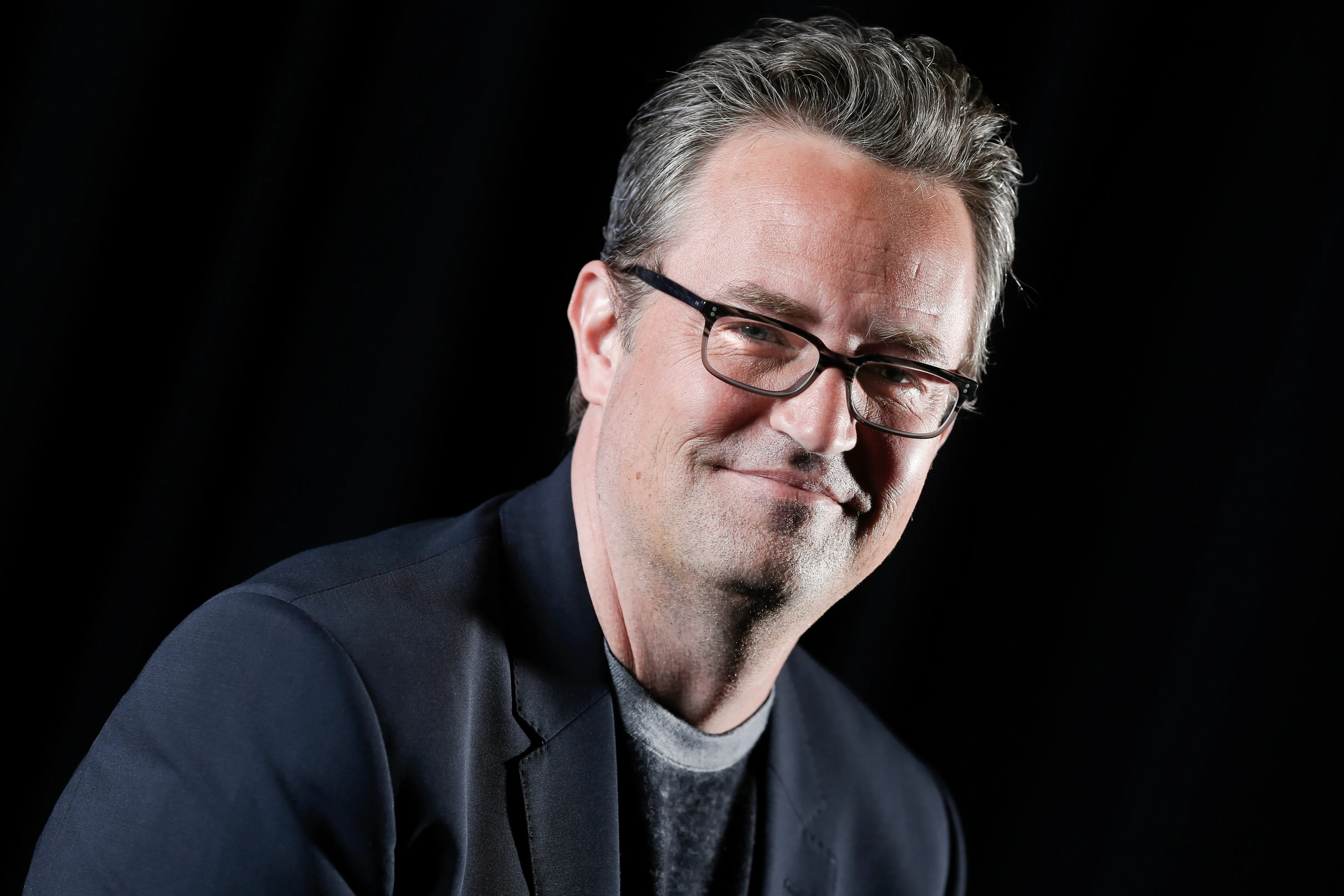 Matthew Perry smiles in glasses in a portrait against a black background.