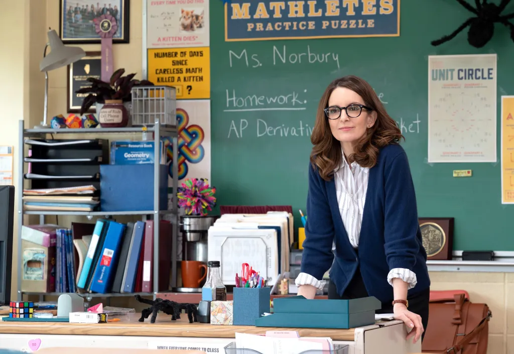 Tina Fey as Ms. Norbury in a classroom, leaning on a desk in front of a blackboard with math problems and an intimidating sign that reads, 