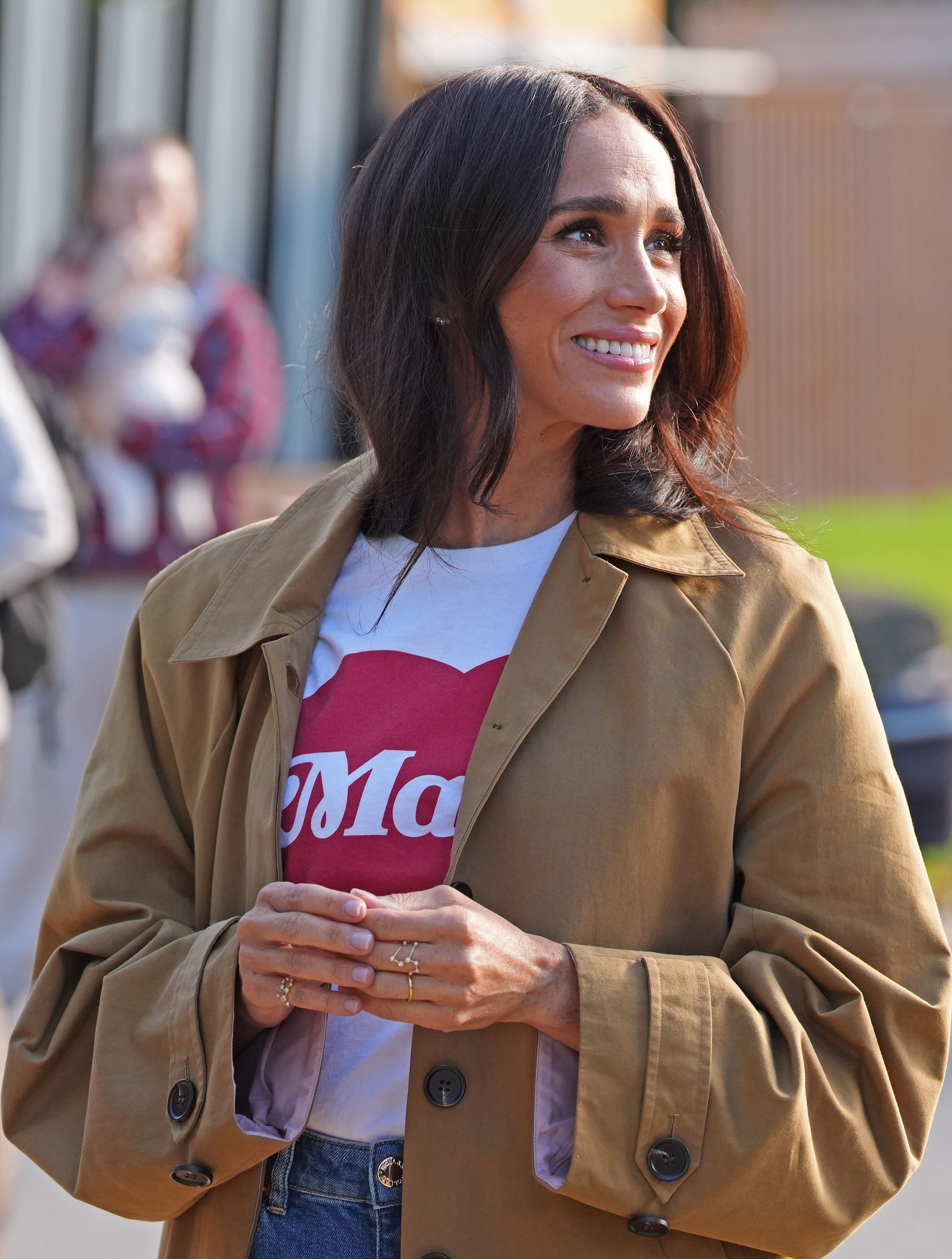 Meghan Markle smiling while wearing a white t-shirt, brown jacket, and jeans.