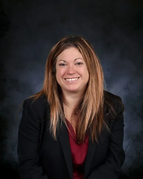 Meghan Manion, a woman with long light brown hair, smiles at the camera while wearing a black blazer and a red top.
