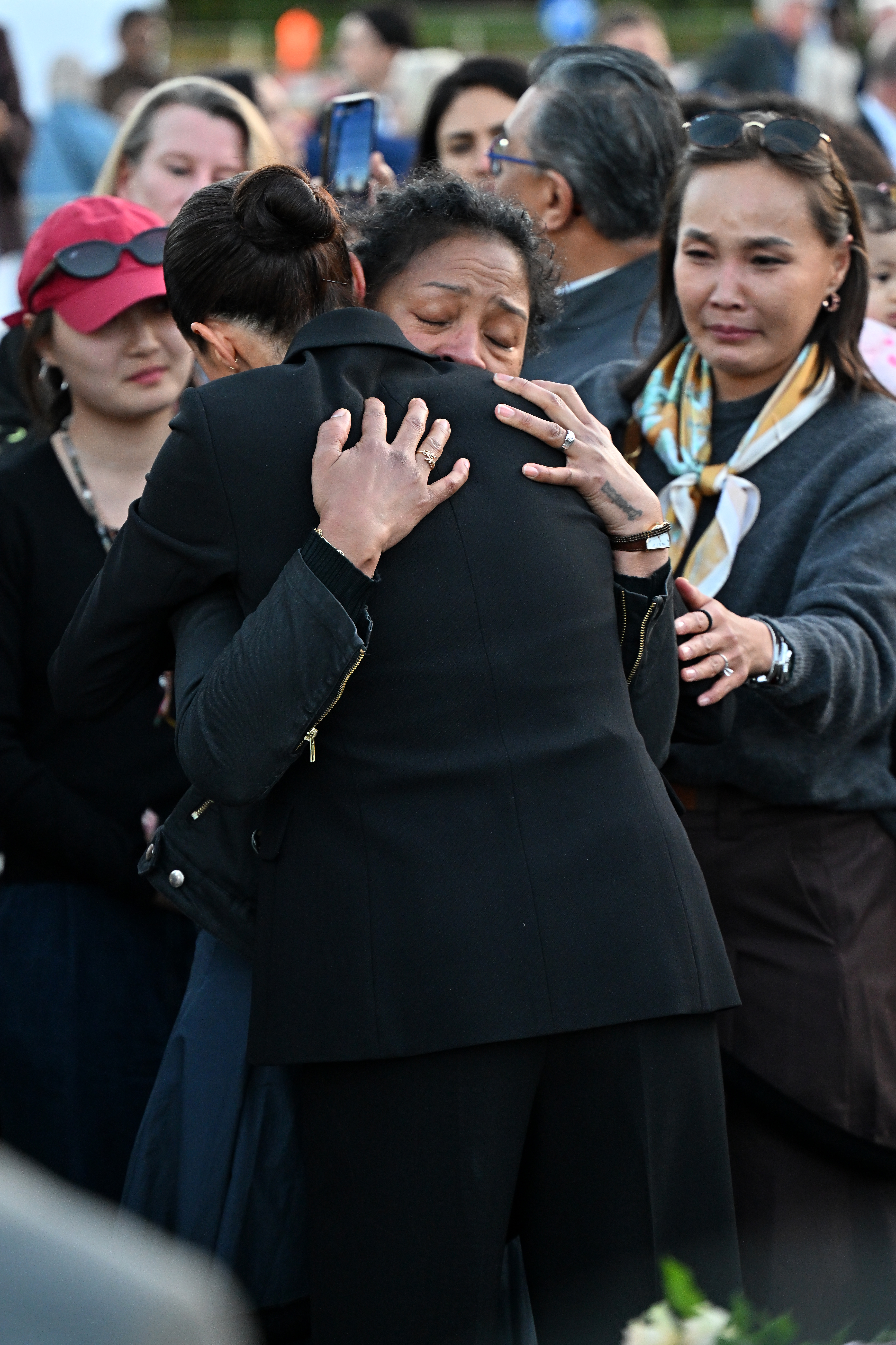 A woman in a black jacket embracing a crying woman, surrounded by onlookers.