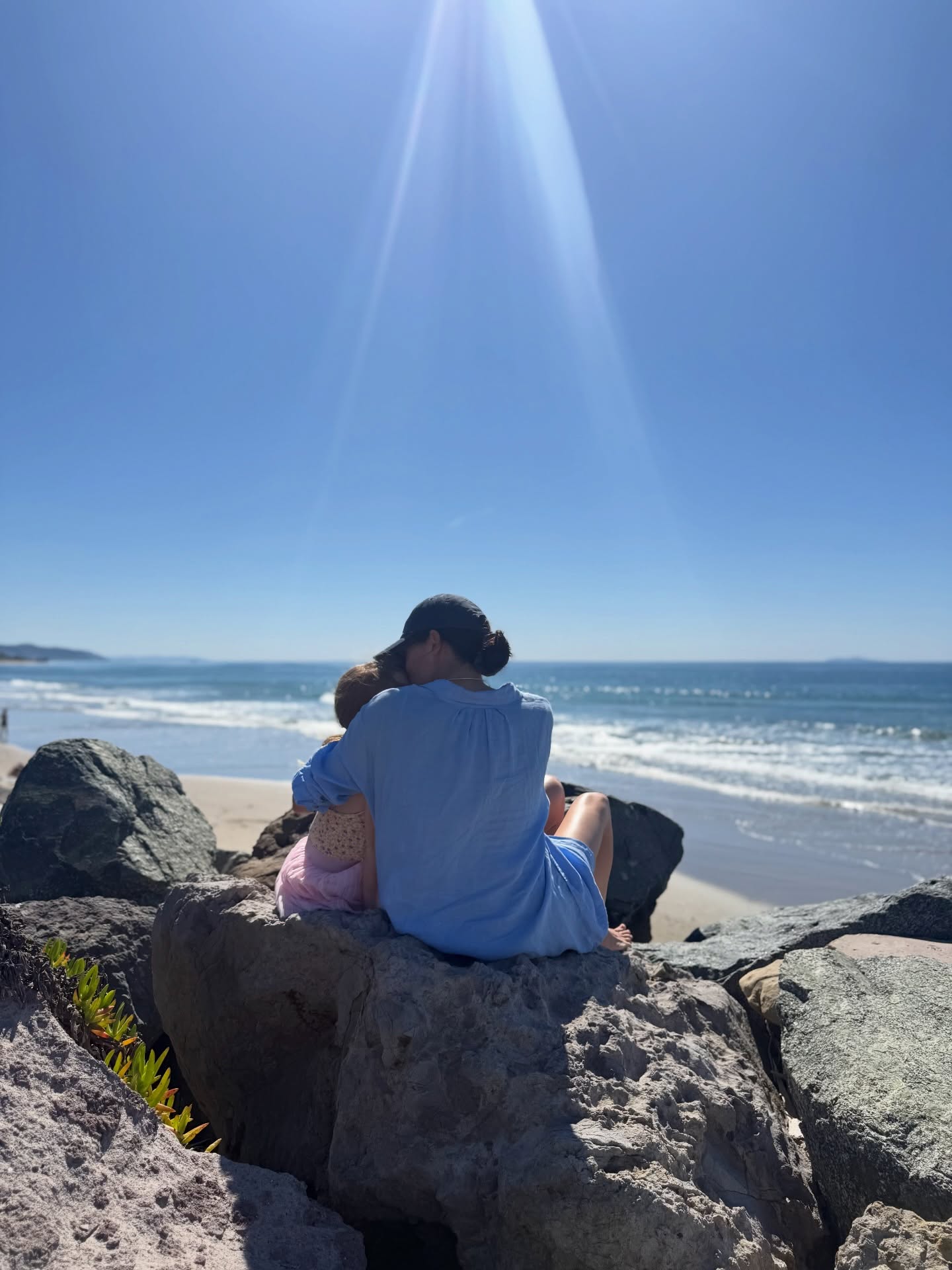Meghan Markle holding Lilibet while sitting on rocks overlooking the ocean.
