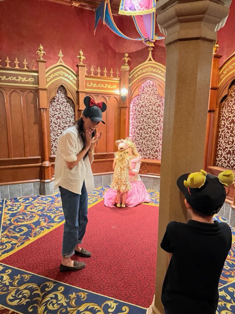 Meghan Markle in a Minnie Mouse hat at Disneyland, smiling while a child in a floral dress approaches Princess Aurora.