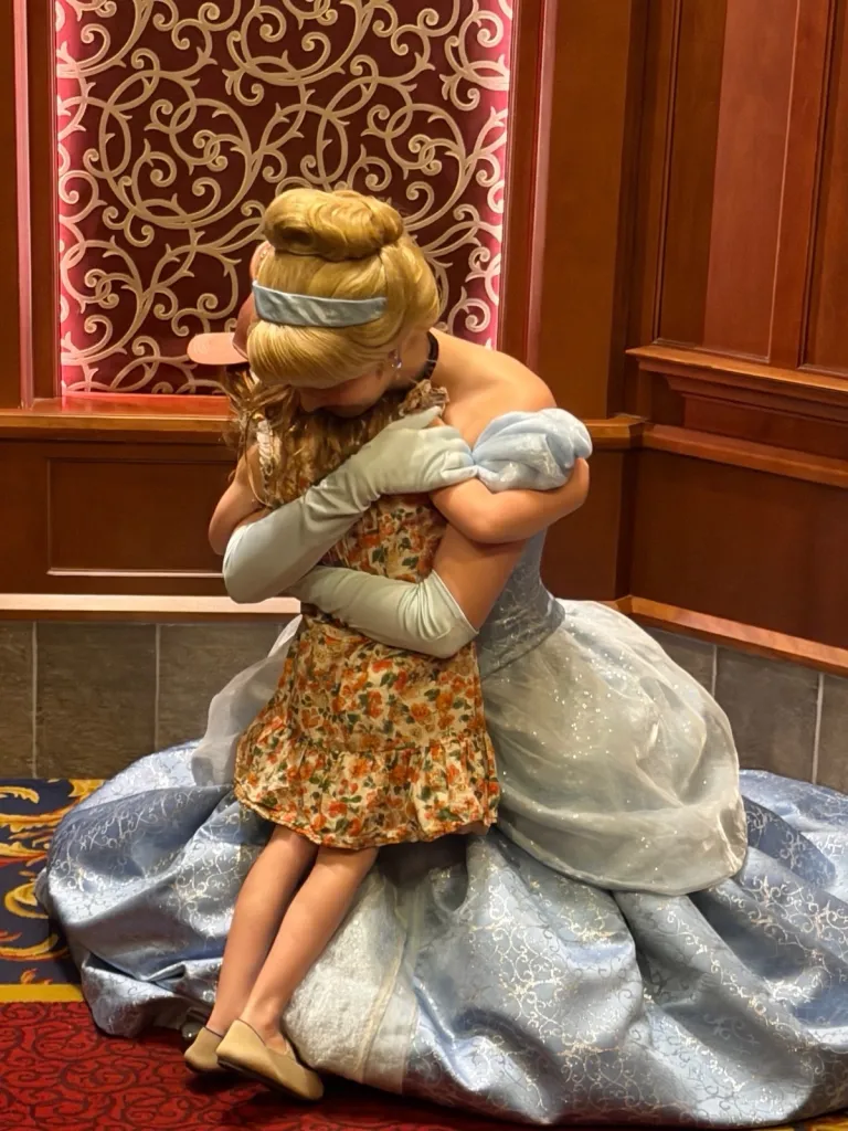 Cinderella character in a blue gown embracing a young child in a floral dress.