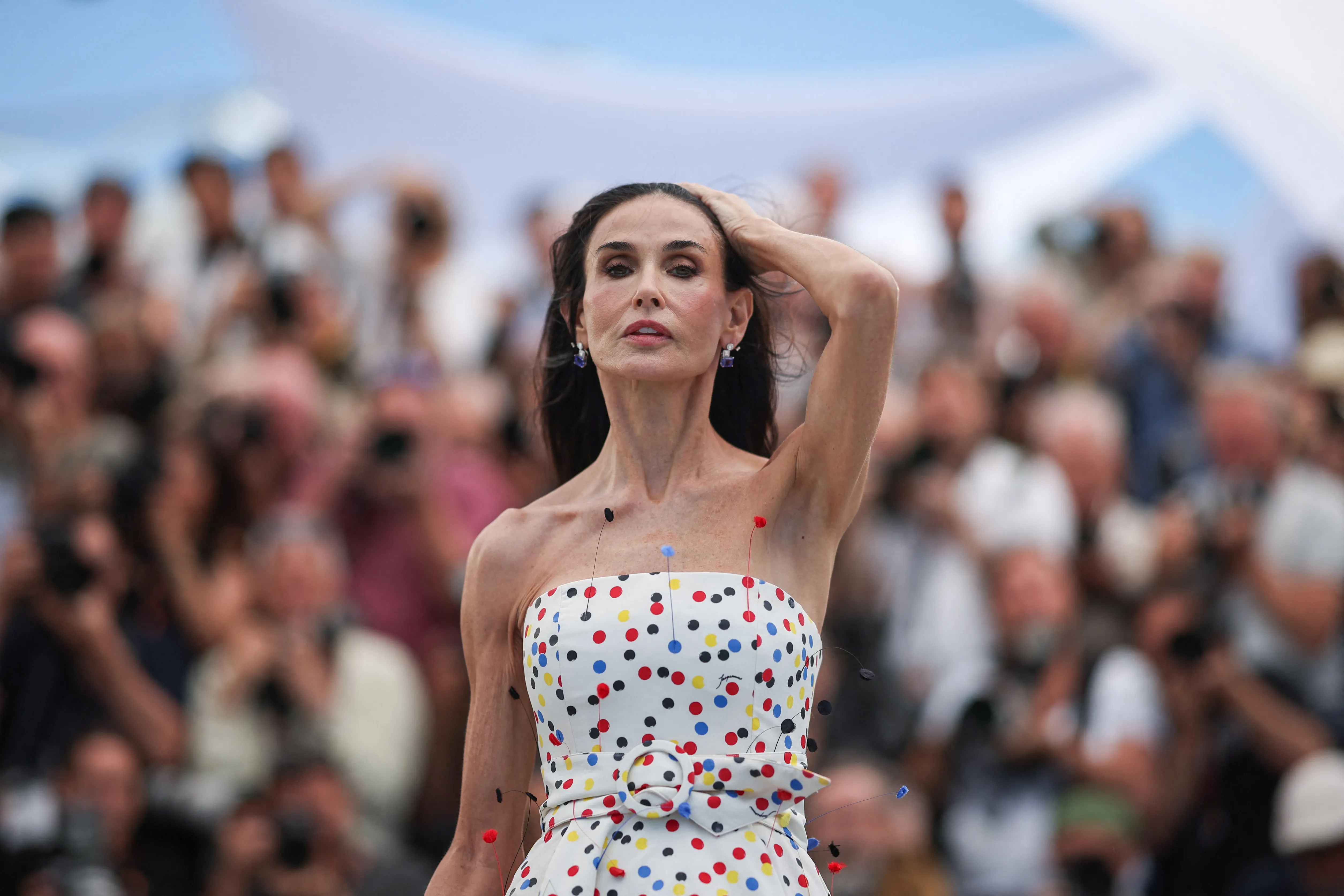 Demi Moore posing with her hand behind her head while wearing a white strapless dress with colorful polka dots, with an audience in the blurred background.