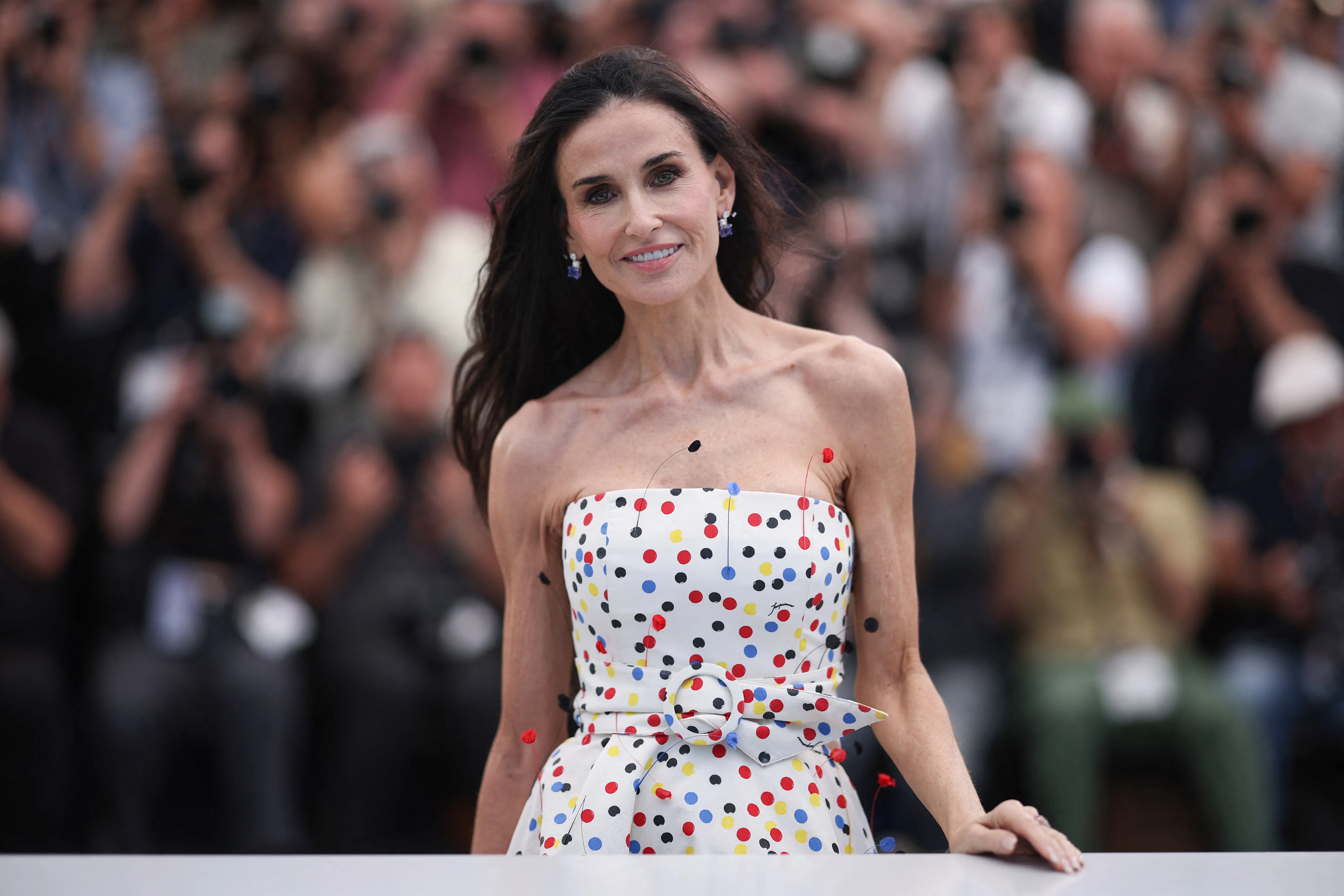 Demi Moore smiling in a white polka dot dress with a large buckle belt, at the Cannes Film Festival.