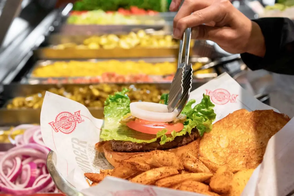 A hand using tongs to place an onion ring on a hamburger with lettuce, tomato, and a side of fries.