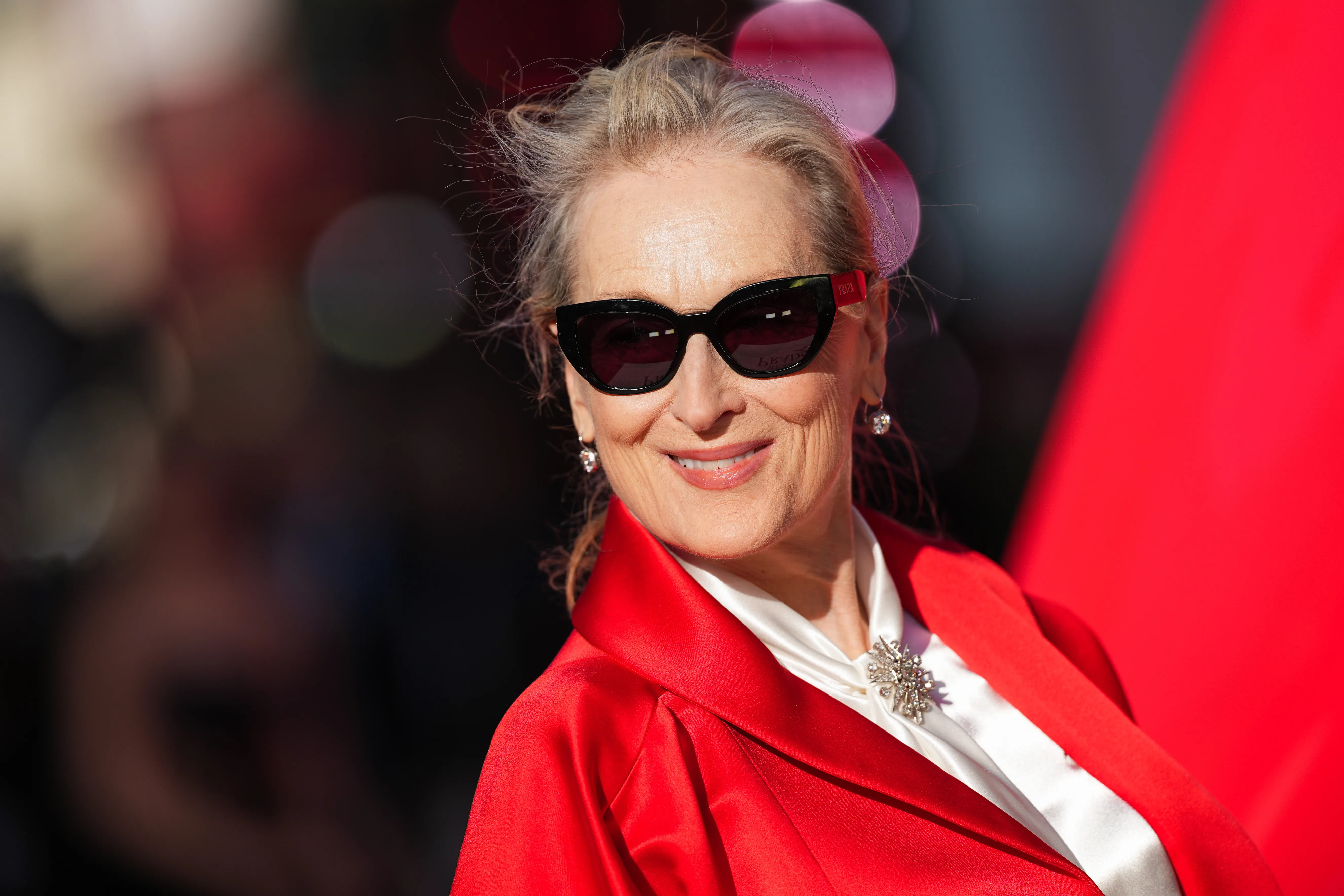 Meryl Streep smiles wearing black sunglasses and a red satin jacket over a white blouse with a decorative pin.