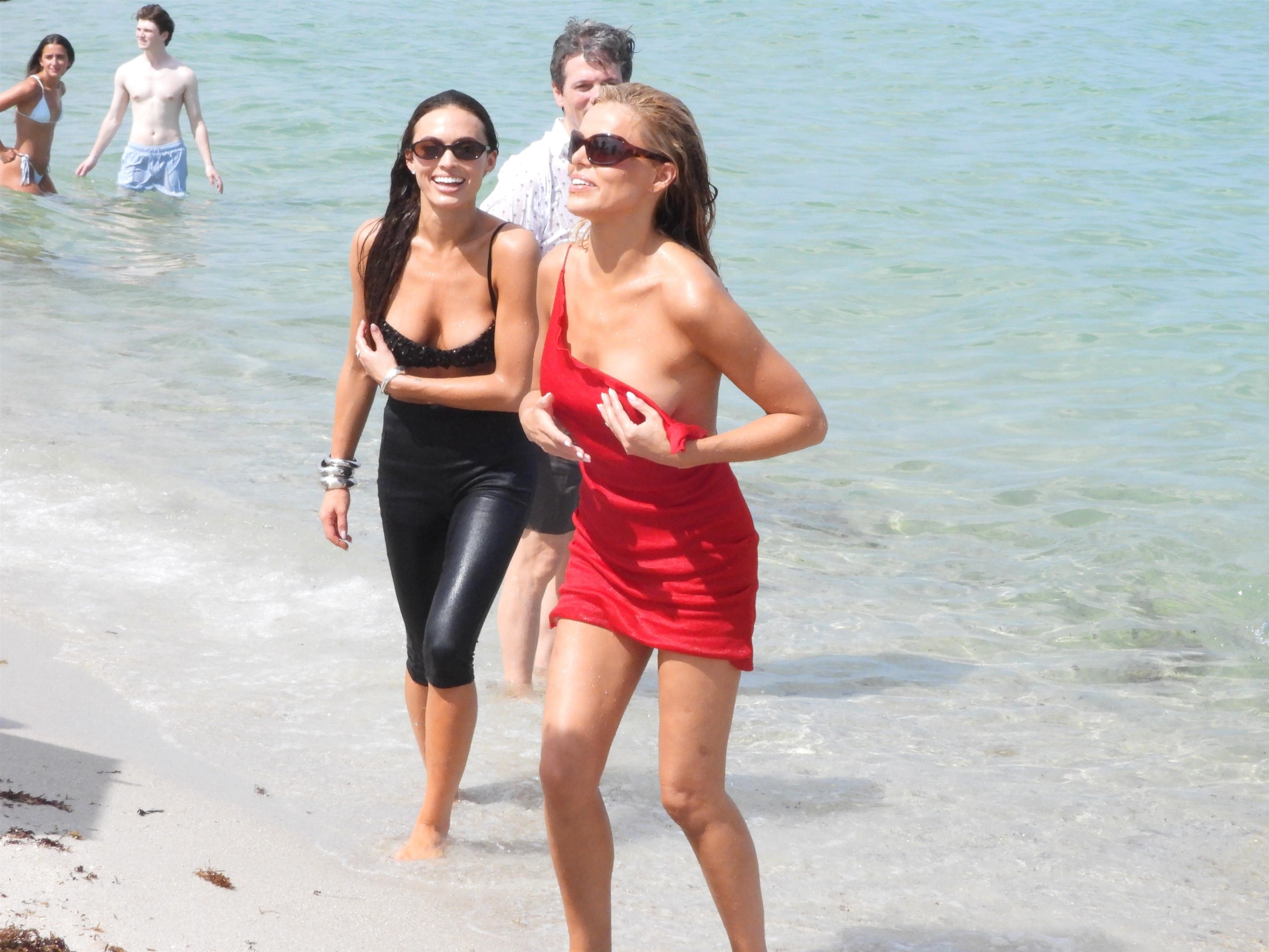 Brooks Nader in a red dress with another woman in a black outfit, both on a Miami beach, with other people in the background.