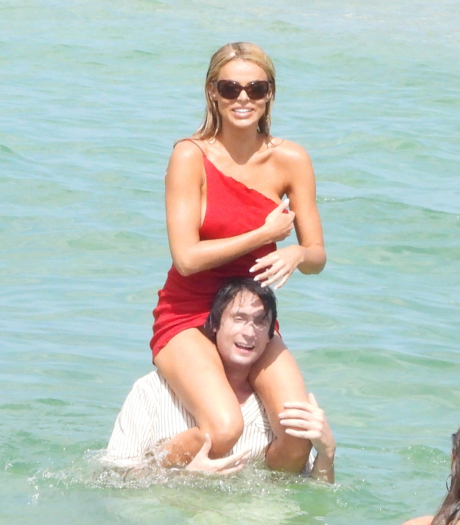 Brooks Nader in a red dress being carried on the shoulders of a man in the ocean.