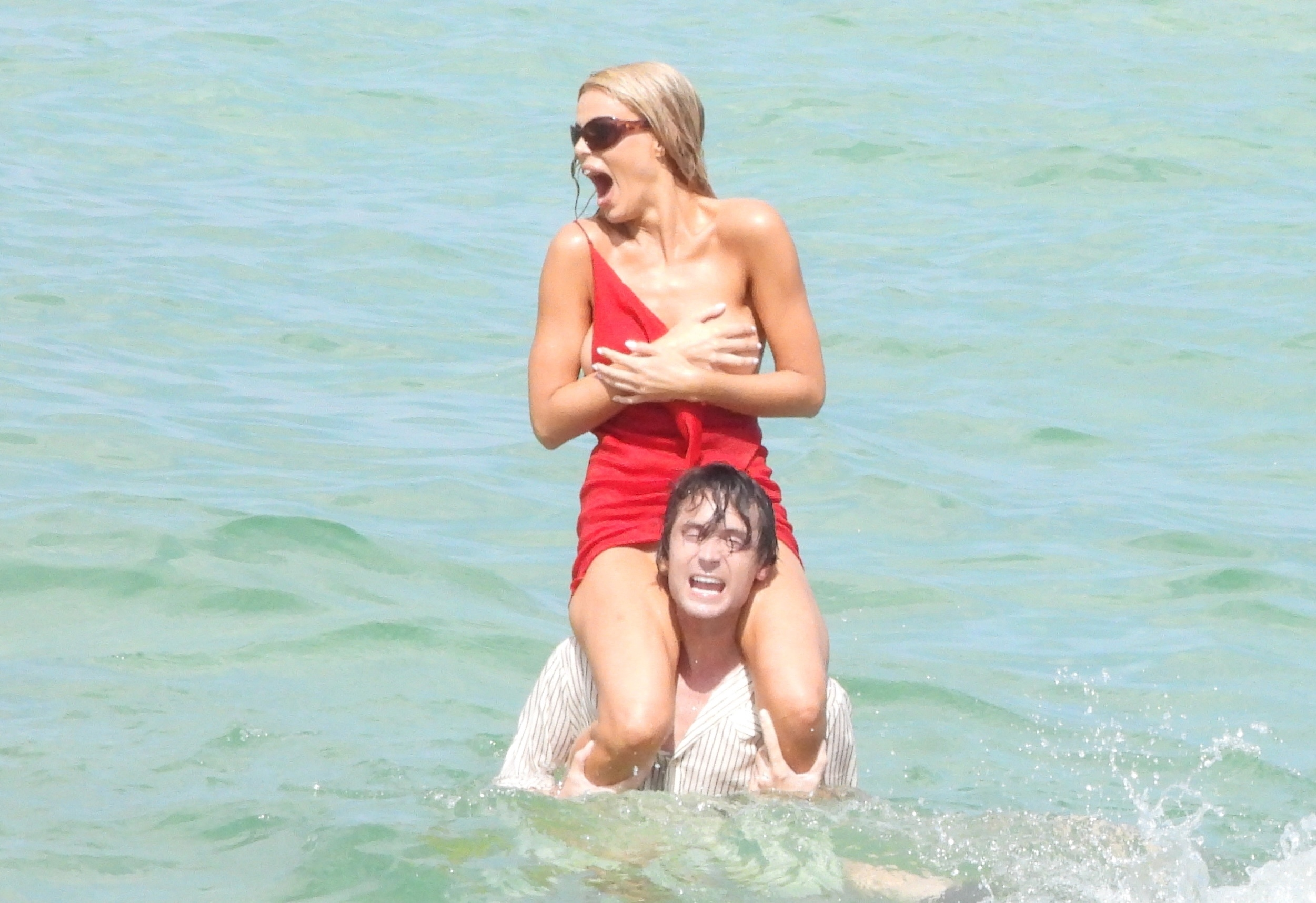 Brooks Nader in a red dress covering herself on a man's shoulders, yelling in the water.