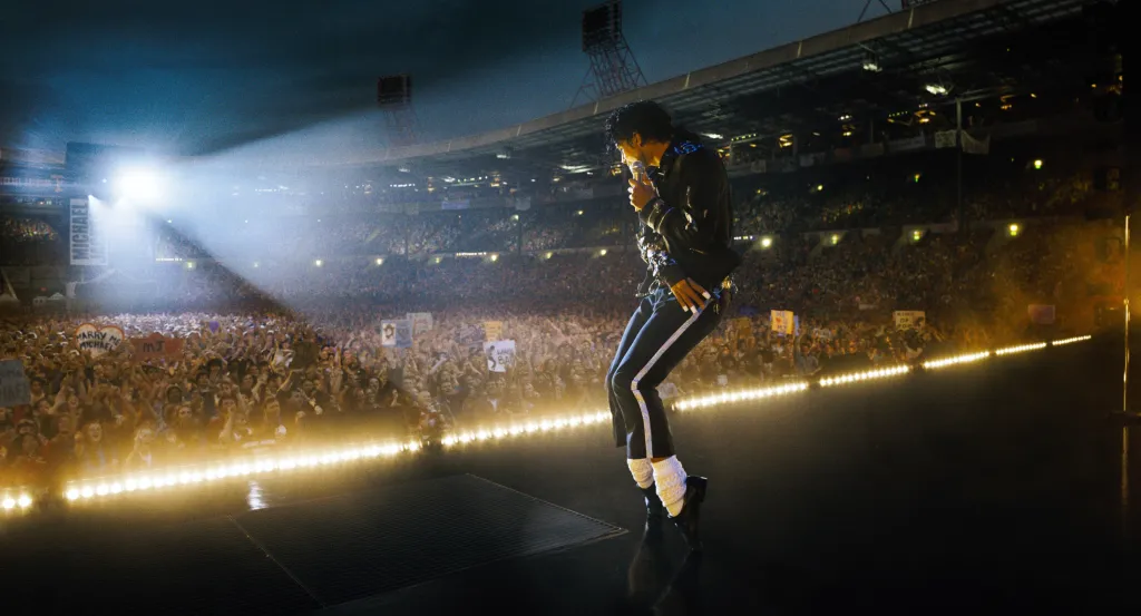 Jaafar Jackson as Michael Jackson on stage in a black jacket and pants, performing for a large crowd in a stadium.