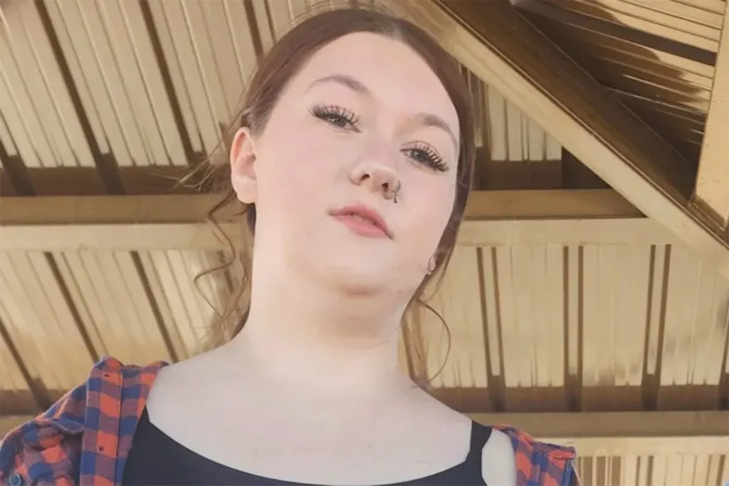 Photo of a young woman looking directly at the camera, with a nose piercing, wearing a black top and a plaid shirt.