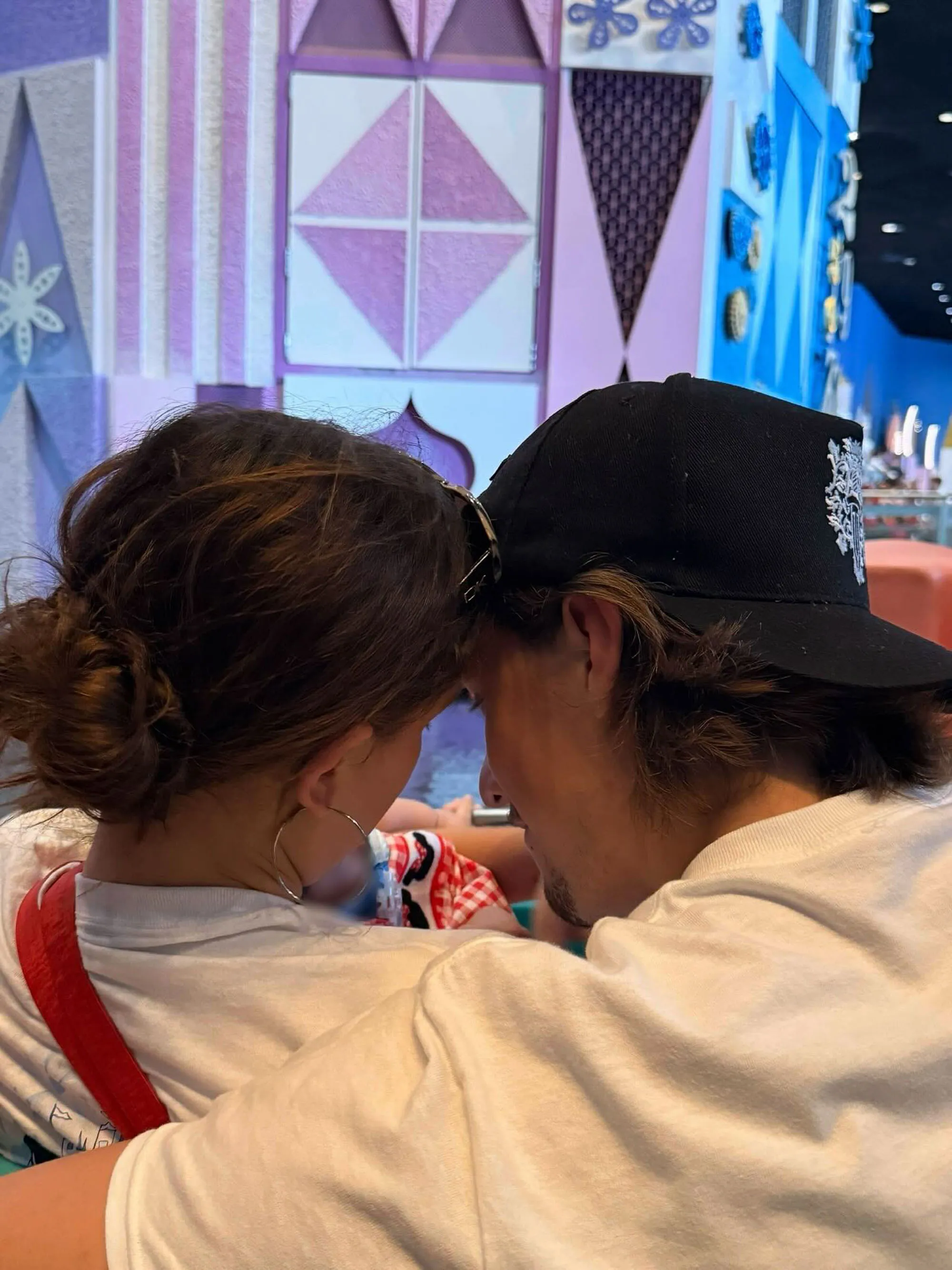 Millie Bobby Brown and Jake Bongiovi with their baby daughter, with their heads together and baby lying between them, against a colorful cartoon backdrop.