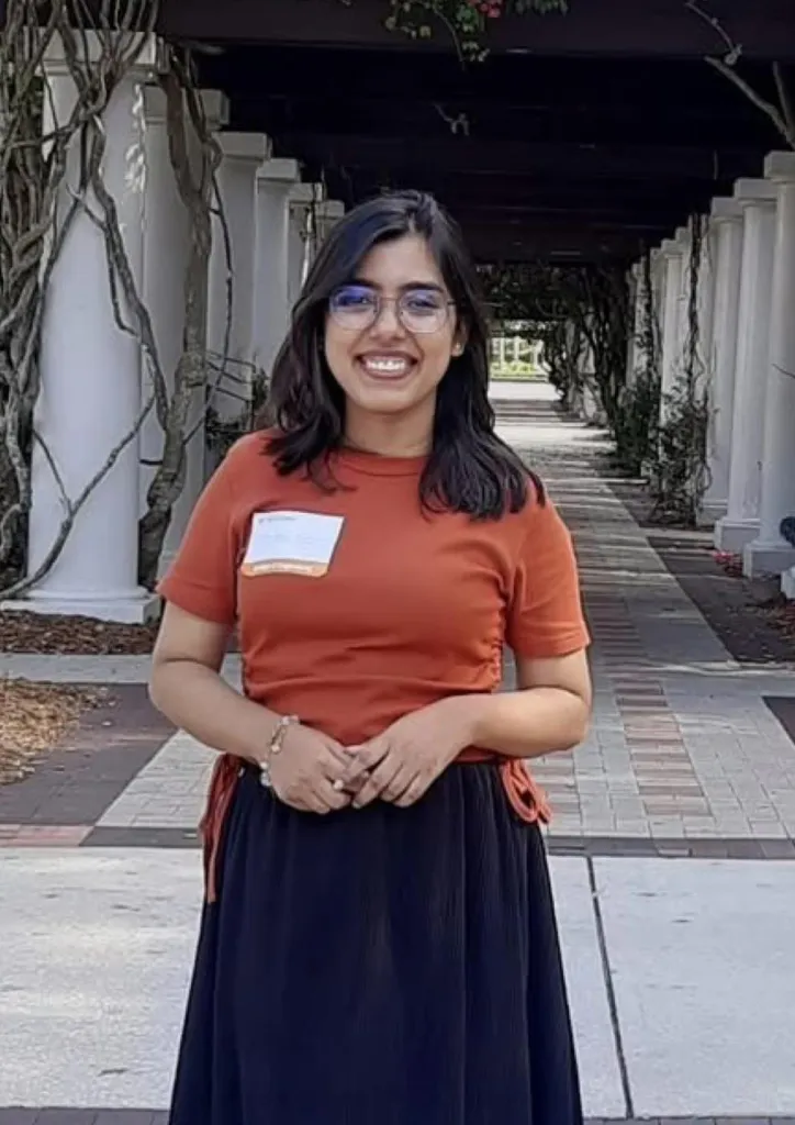 A young woman with dark hair and glasses smiles at the camera, wearing an orange shirt and black skirt.