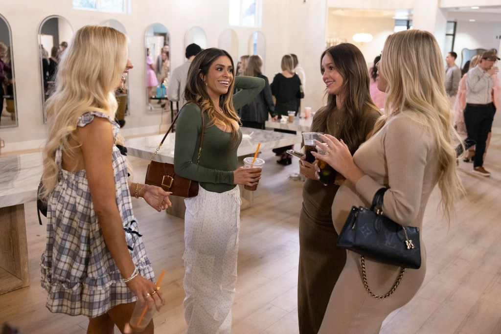 Four women talking in a room, two on the left holding drinks, and two on the right holding drinks and one also holding a phone.