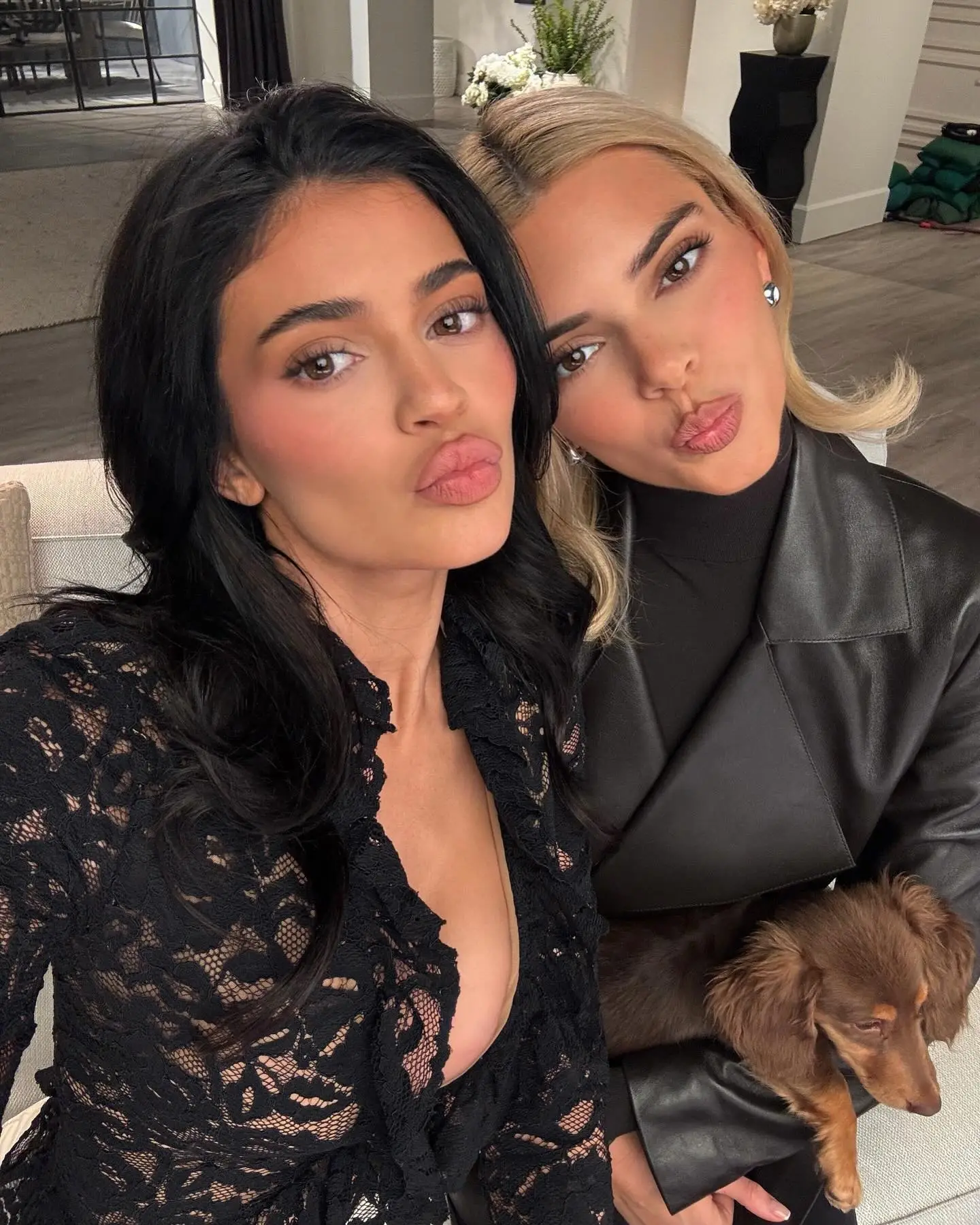 Kylie Jenner in black lace and Kendall Jenner in a black leather jacket with a small dog, both making kissing faces.