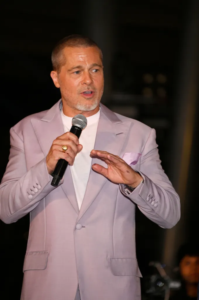 Brad Pitt speaking into a microphone, wearing a light purple suit jacket with a white t-shirt underneath.