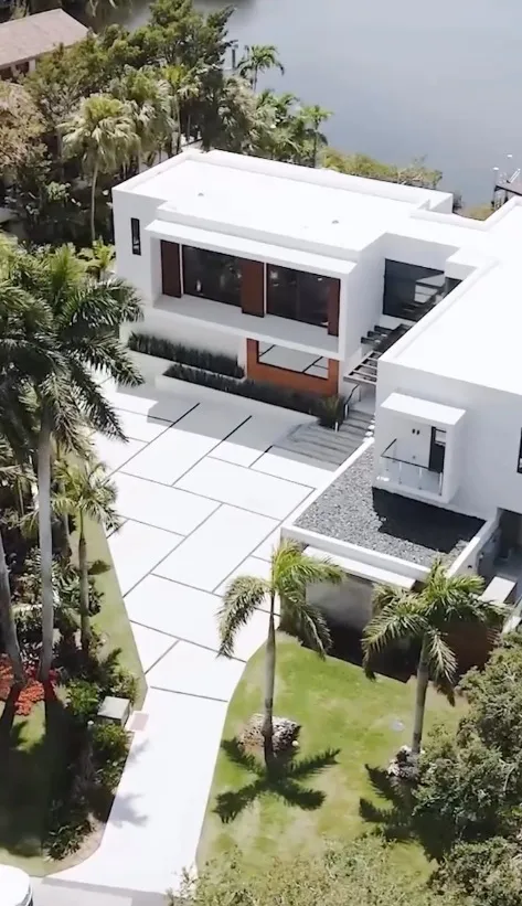 Aerial view of a modern white house with a paved driveway and palm trees, with a body of water in the background.
