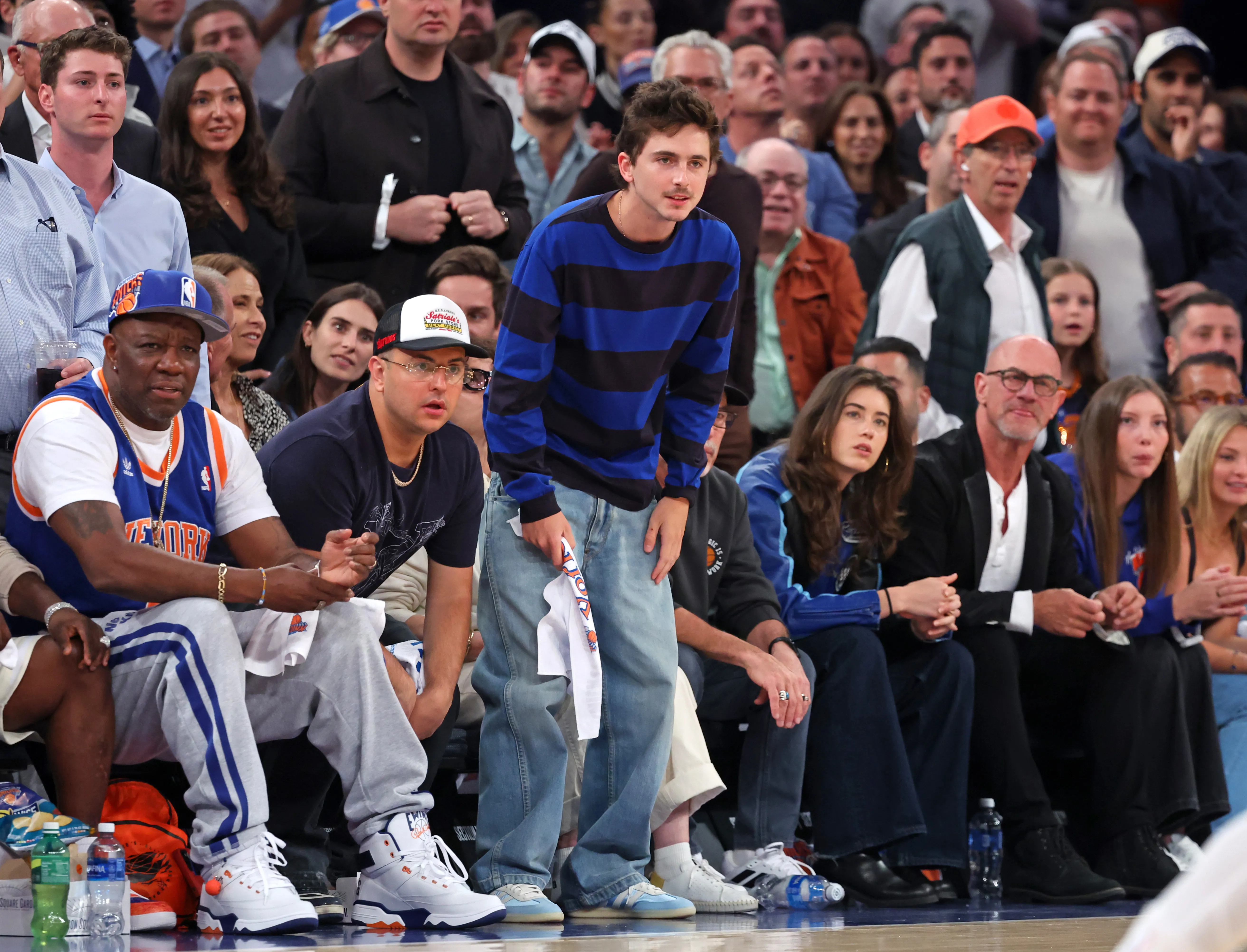Actor Timothee Chalamet watches a basketball game from the sidelines, surrounded by other spectators.
