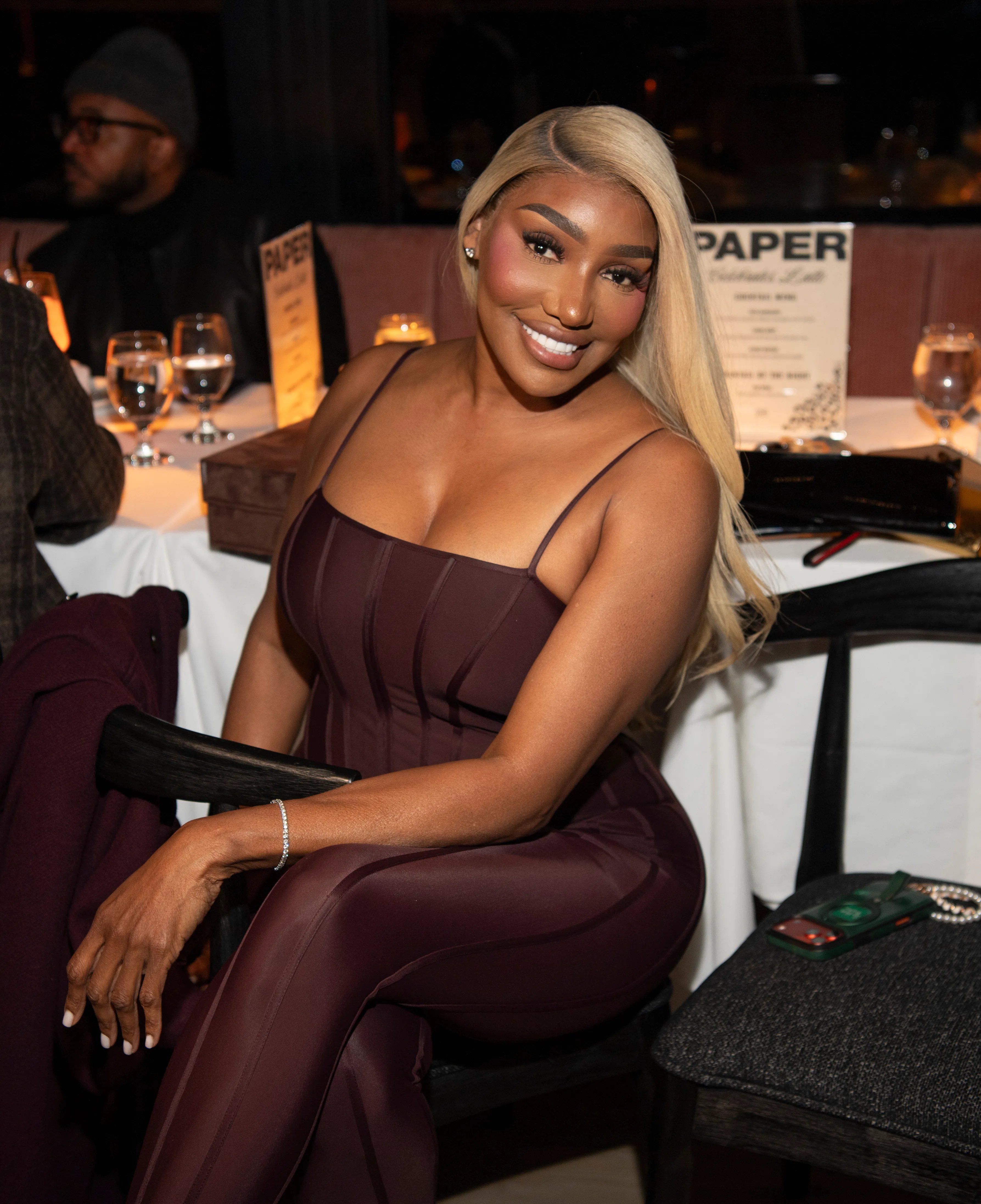 Nene Leakes smiling, wearing a dark burgundy jumpsuit, seated at a restaurant table.